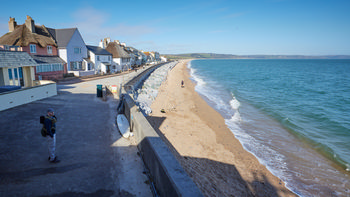Slapton sands sea wall This landscape photograph shows the Slapton Sands sea wall running alongside the beach and coastal village in Devon, England, United Kingdom. Taken in the early afternoon during the spring season, the scene features clear blue skies and sunlight illuminating the picturesque beach and the gentle waves of the English Channel. The image captures people present along the coast, including Jamie Weinbren, who is standing on the walkway overlooking the sea wall. Thatched and traditional houses line the road adjacent to Slapton Sands, with fishing activity visible on the sand and distant green hills further enhancing the coastal environment. This photograph showcases the relationship between the coastal protection of Slapton Sands and the beachside community, highlighting a popular location in Devon for recreation and scenic views.